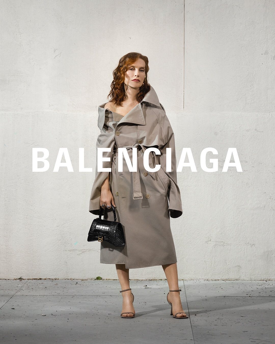 Adult woman poses in an asymmetrical beige trench coat in front of a textured white wall while holding a black reptile purse.