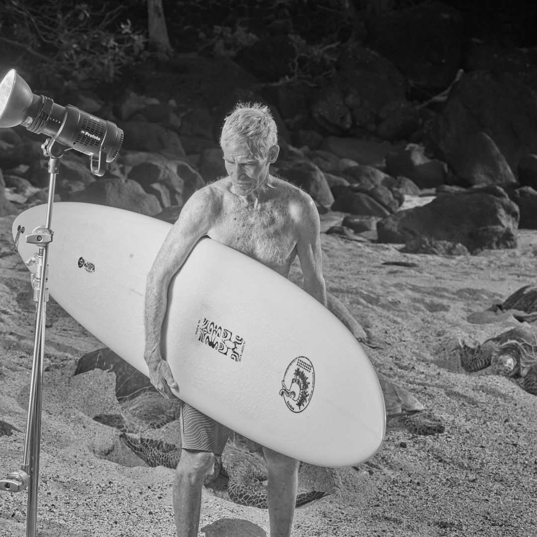 Black and white shot of a mature adult man holding a surfboard on a sandy beach next to a professional photography light stand.