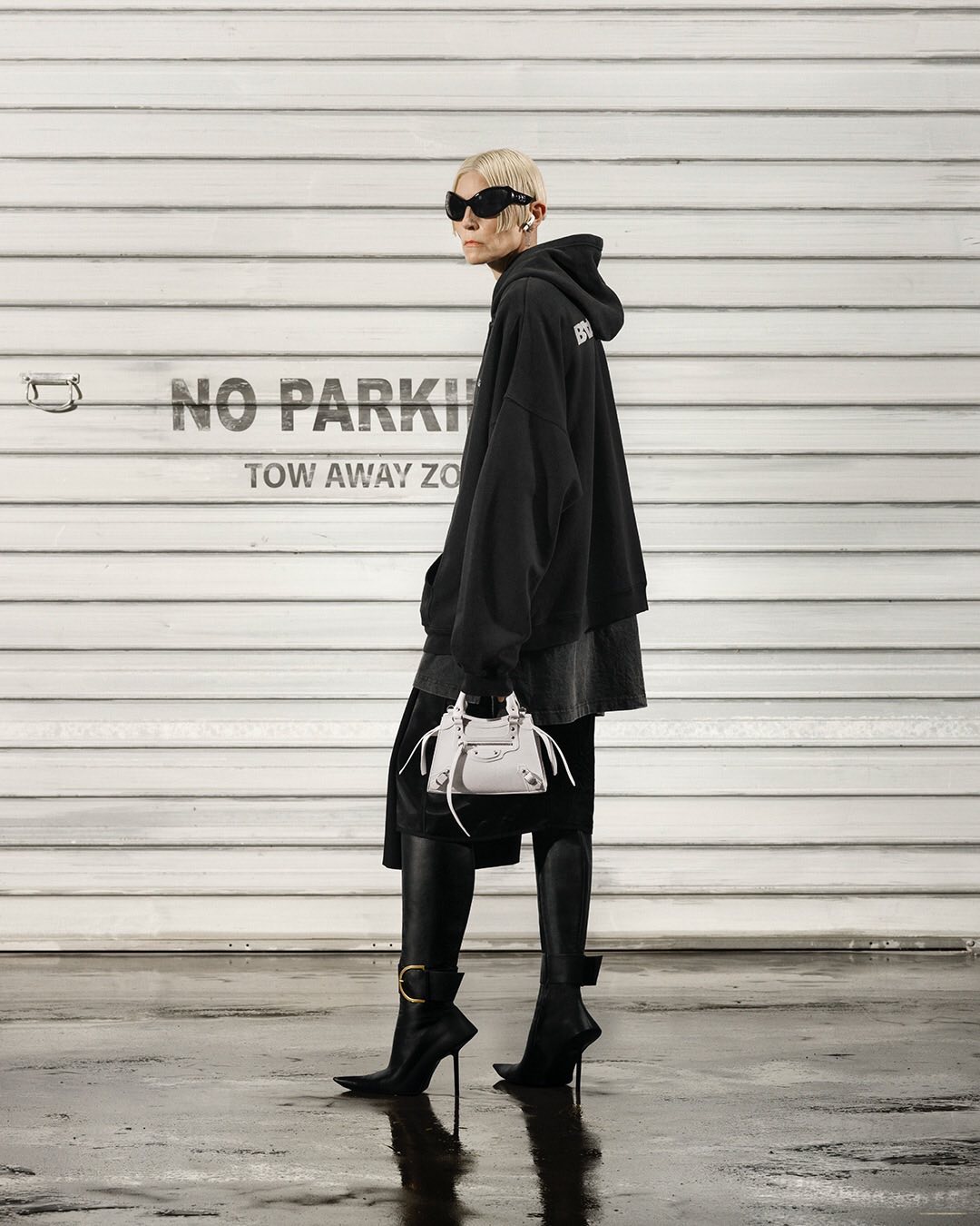 Woman in oversized hoodie, sunglasses, and high heel boots walks past a white garage door holding a small white handbag.
