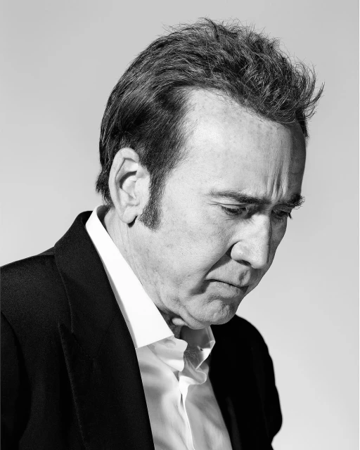 Black and white portrait of Nicolas Cage wearing an open white collared shirt and a black suit jacket against a light neutral background.