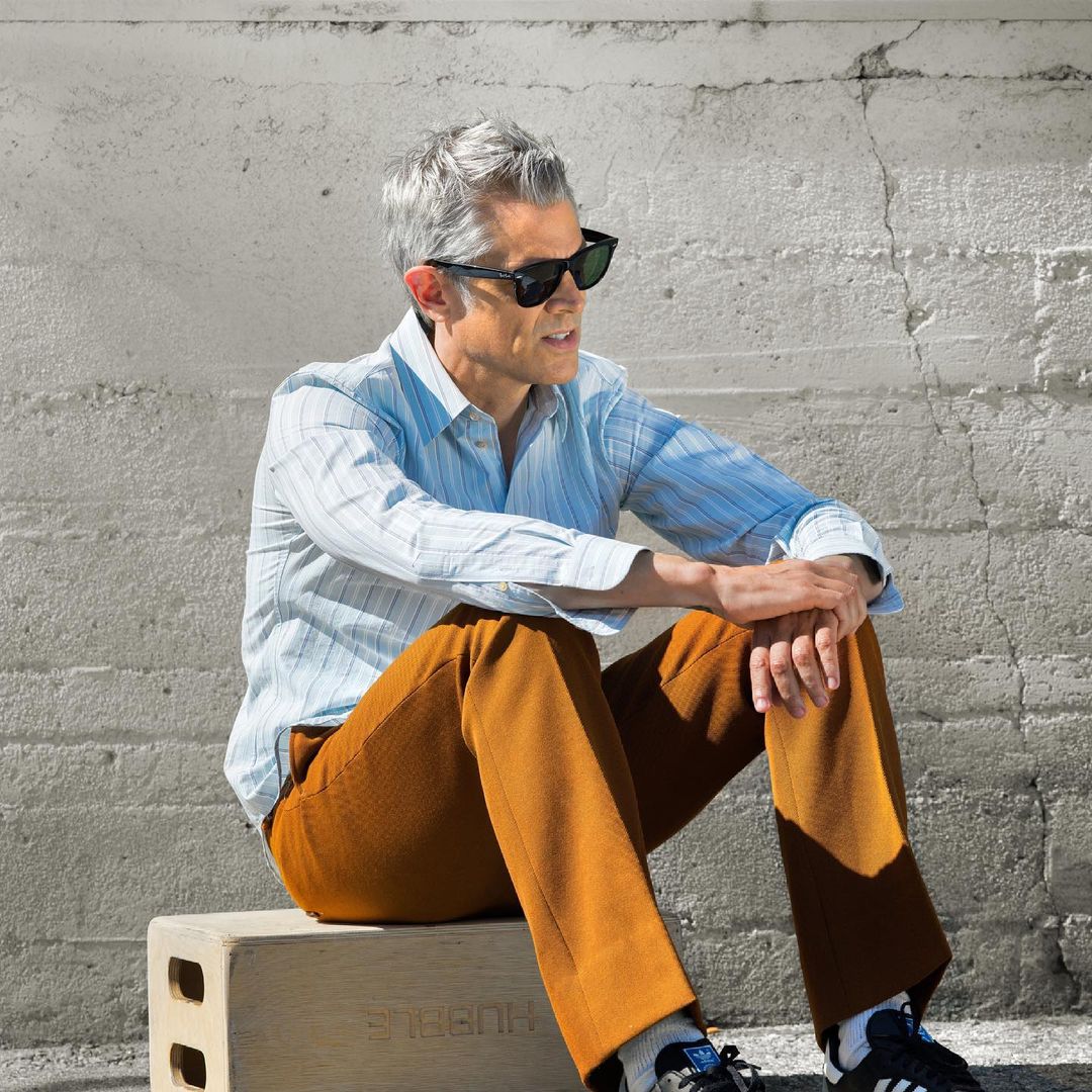 Stylish adult man with gray hair and sunglasses sits on a block outside, wearing a striped shirt and brown pants, creating a fashion portrait.