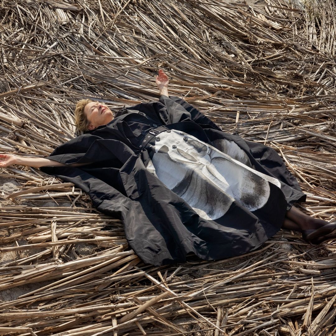 Adult woman in stylish black dress with abstract print lies on a bed of reeds outdoors, bathed in sunlight.