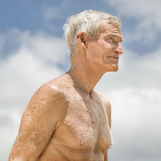 Elderly man with gray hair poses shirtless in a side-view, set against a sky with soft clouds. Natural light illuminates his features.
