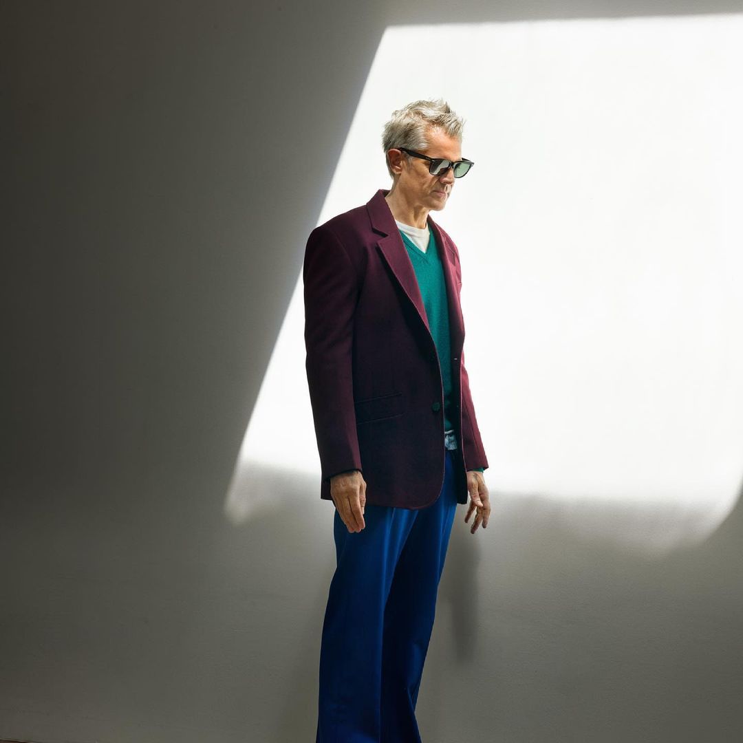 Mature man in burgundy jacket and blue pants, posing in a studio setting, bathed in dramatic window light, showcasing a modern and stylish look.
