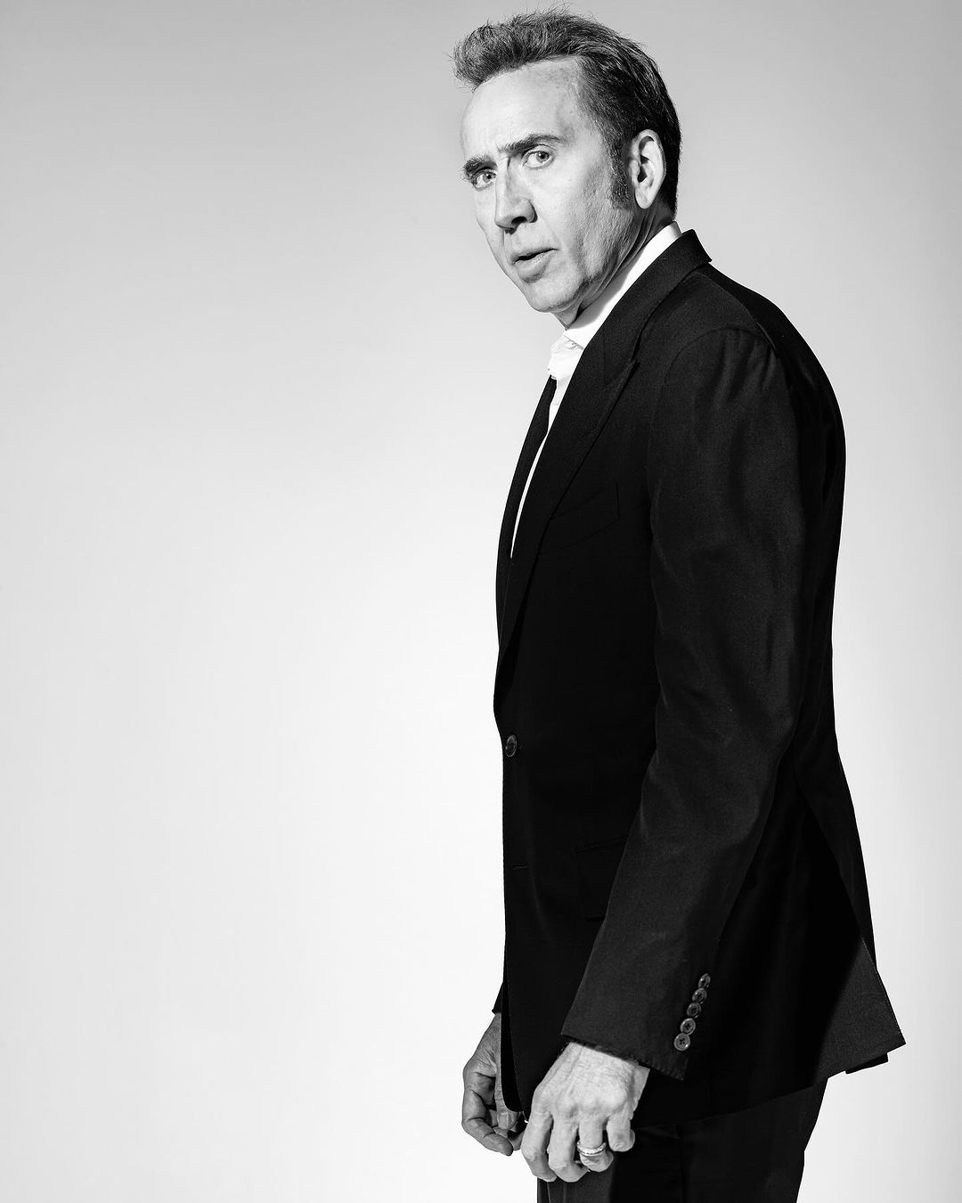 Black and white portrait of Nicolas Cage in a suit looking over his shoulder against a bright white seamless backdrop in a photo studio.