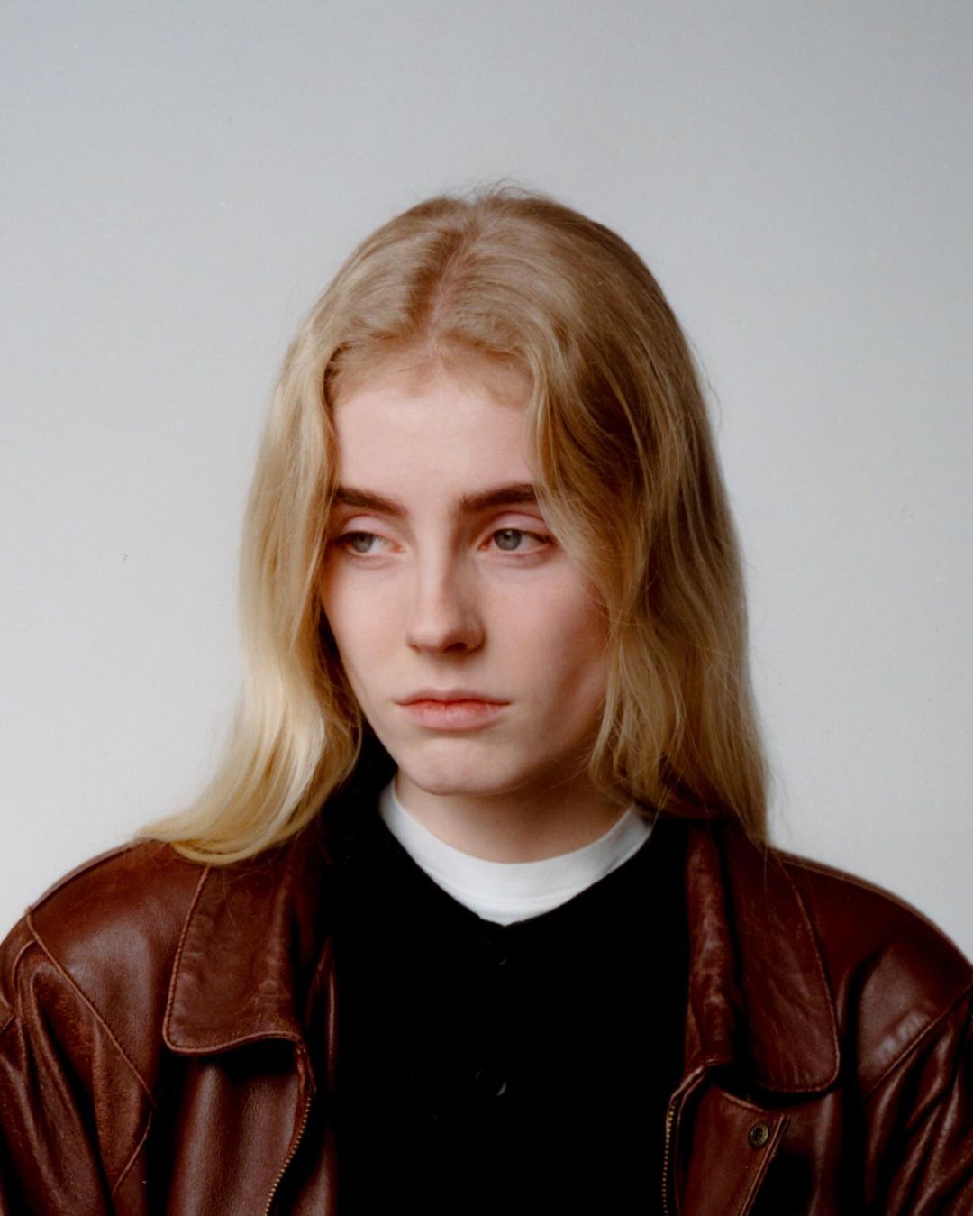Portrait of a young adult woman with long blonde hair wearing a brown leather jacket against a neutral grey background in a studio.