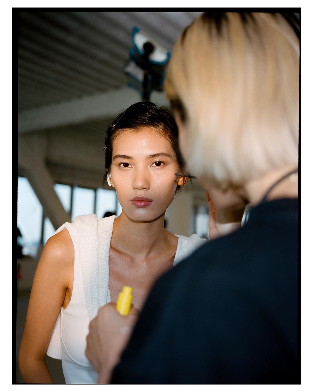 Close-up portrait of a woman with short dark hair getting her makeup done by a makeup artist in a bright studio setting.