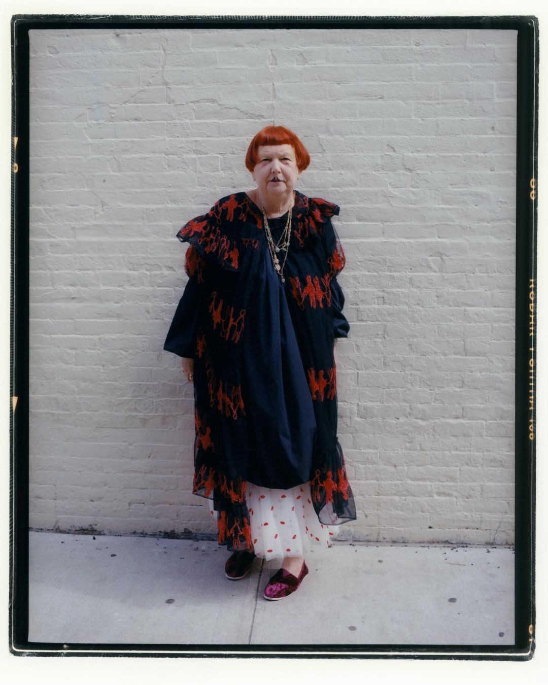 Elderly woman with orange hair standing against a white brick wall, wearing patterned clothing, jewelry, and dark slip-on shoes.
