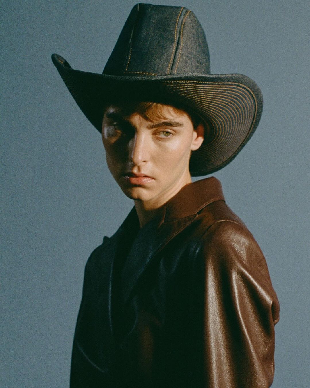 Portrait of a person wearing a denim cowboy hat and a leather coat, posing in a studio with a soft blue backdrop.