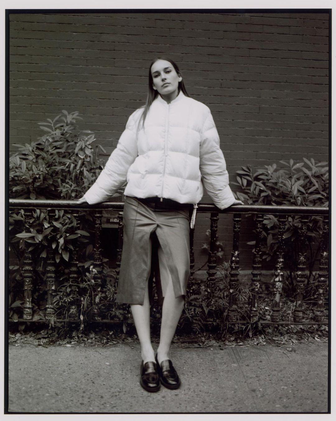 A slender woman in a white puffer jacket and skirt poses in front of a brick wall, leaning on a black railing in an urban setting.