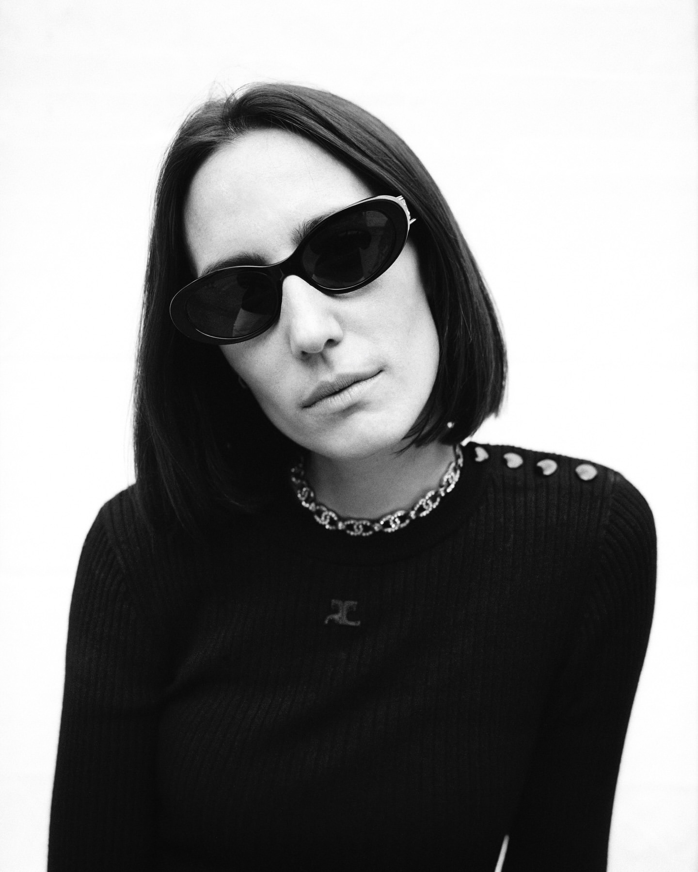 Black and white portrait of a woman with sunglasses wearing a black sweater and silver necklace against a plain white background.