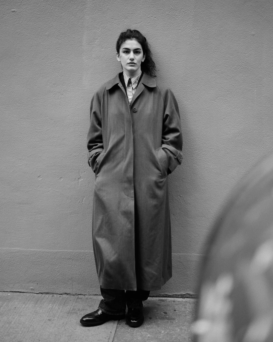 Monochrome portrait of a woman in a long coat standing against a wall, with focus on her expression and the coat's texture.