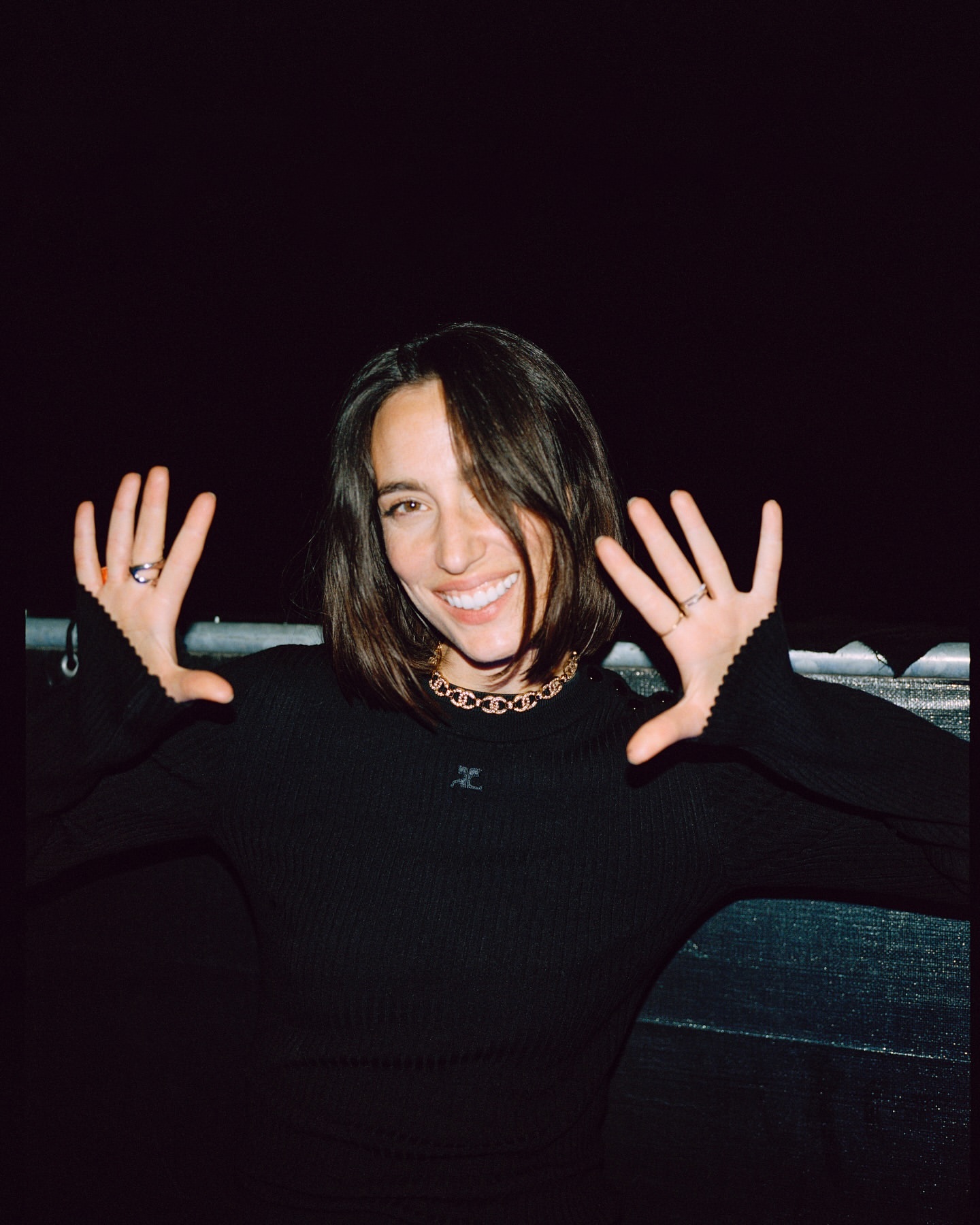 Smiling brunette woman in black top poses with hands up indoors. Close-up captures her happy expression and simple gold necklace.