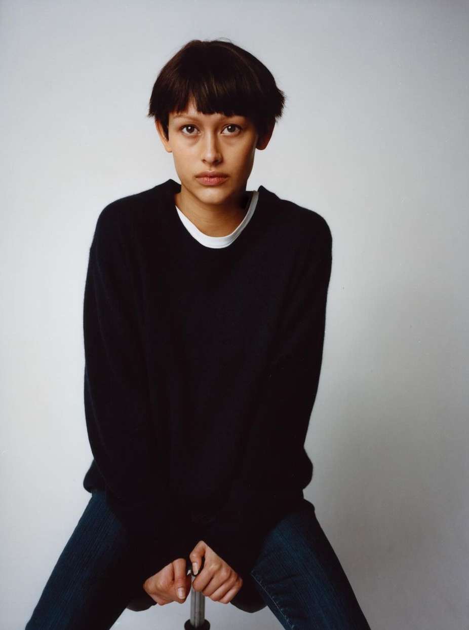 Portrait of a young adult sitting on a rolling stool in front of a solid white background, wearing a dark sweater and jeans.
