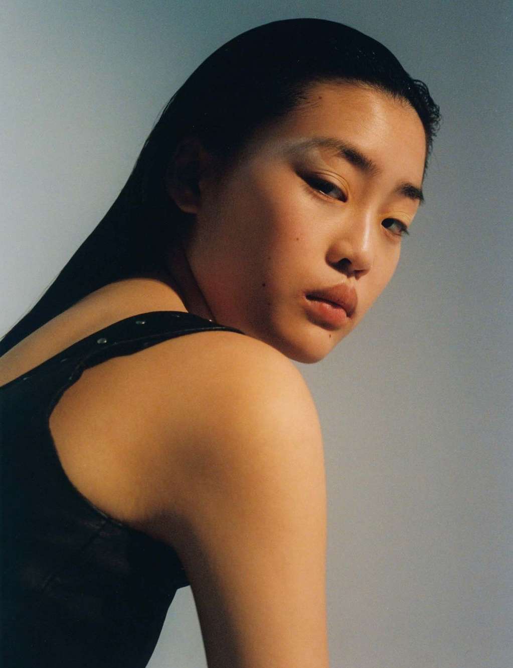 Portrait of a young Asian woman with dark hair, wearing a black top against a neutral background, looking over her shoulder.