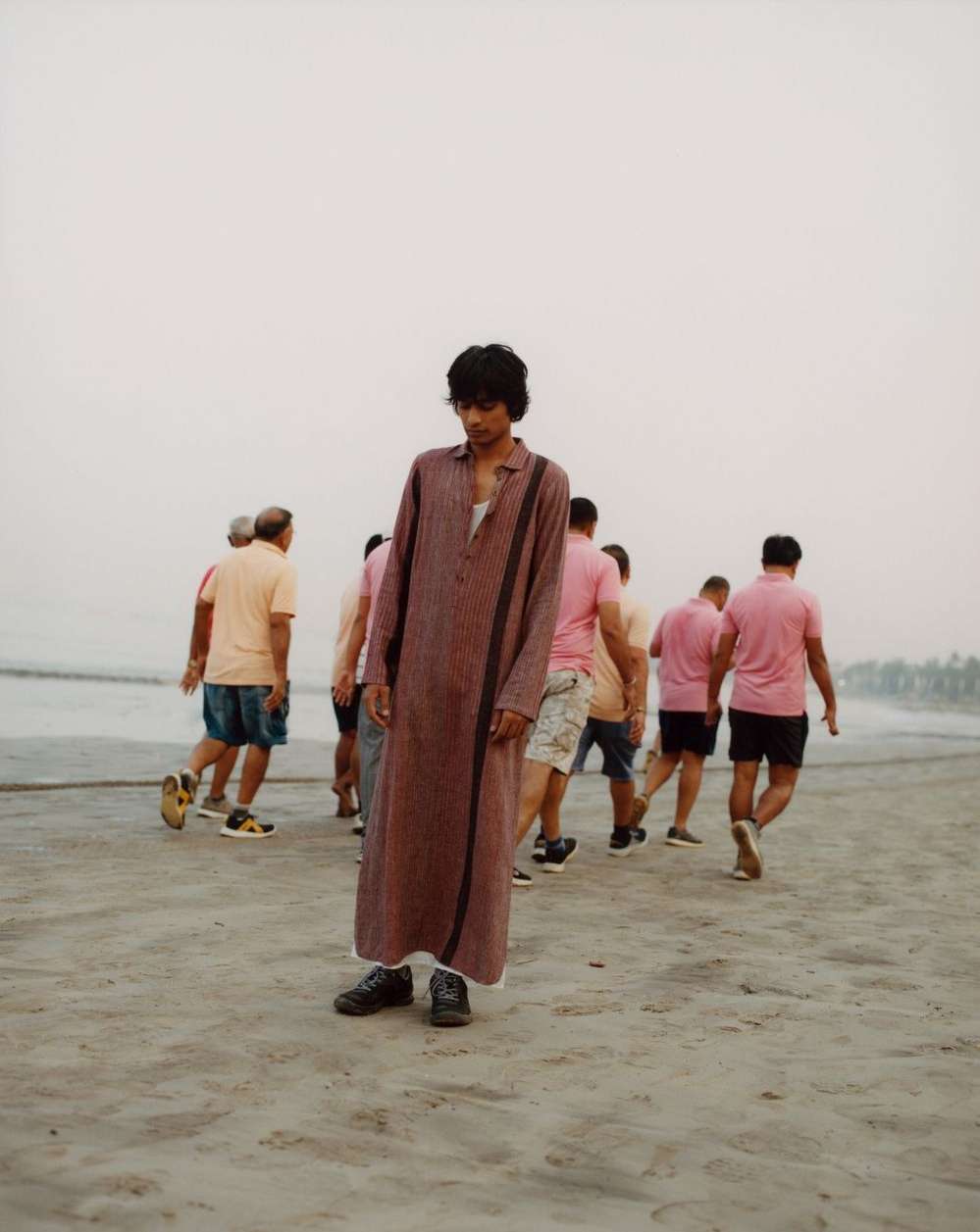 Man in long robe standing on beach with group of men in the background wearing pink shirts and dark shorts on a bright day.