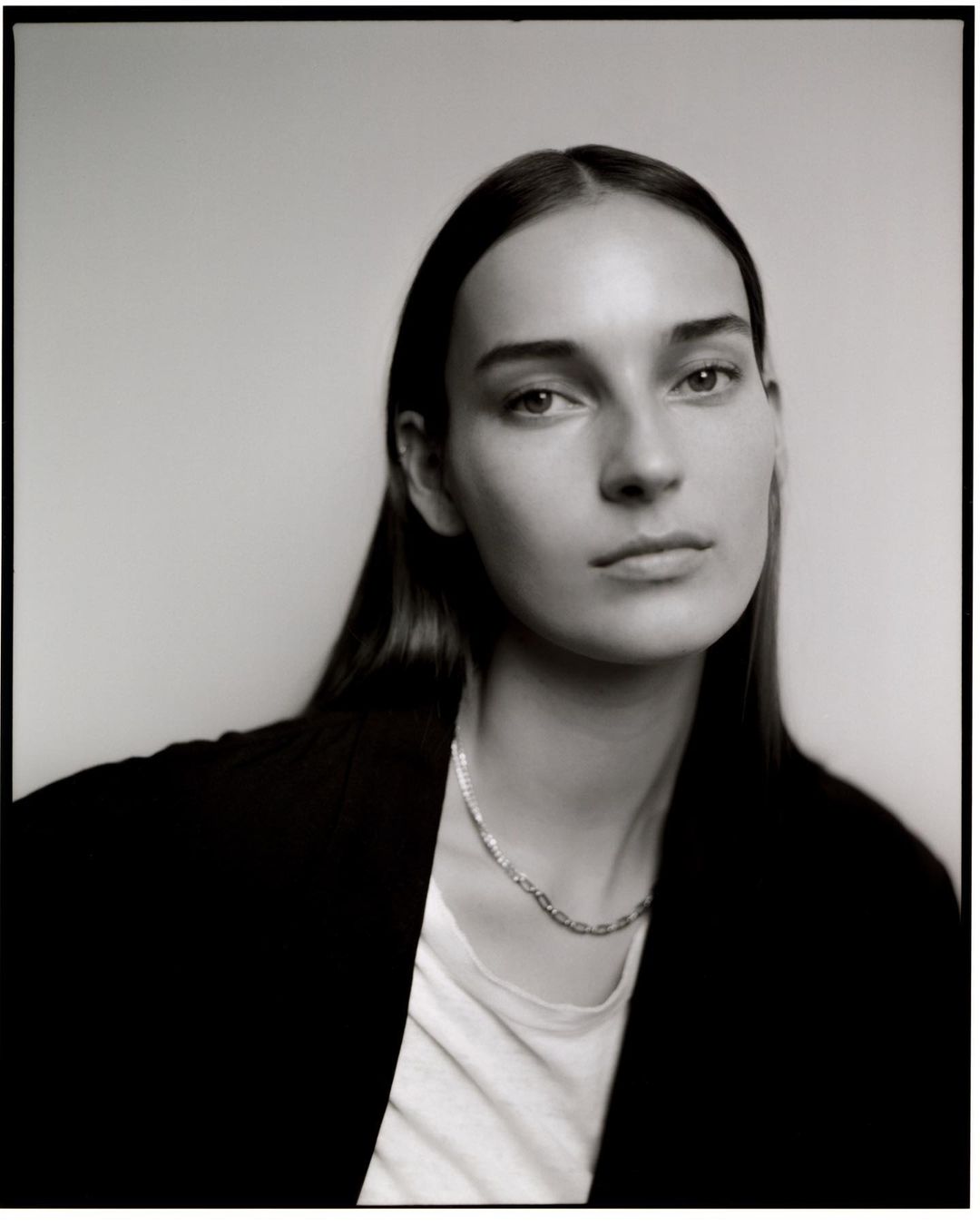 Monochromatic portrait of a young adult female with long hair, wearing a necklace and black jacket against a light background.