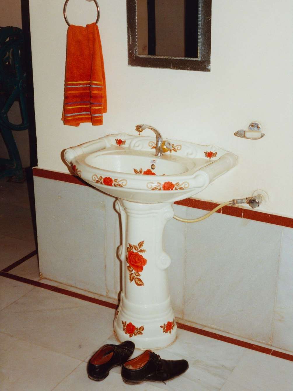 Bathroom interior with a white pedestal sink decorated with red flowers, brown shoes on the floor, orange towel and mirror on the wall.