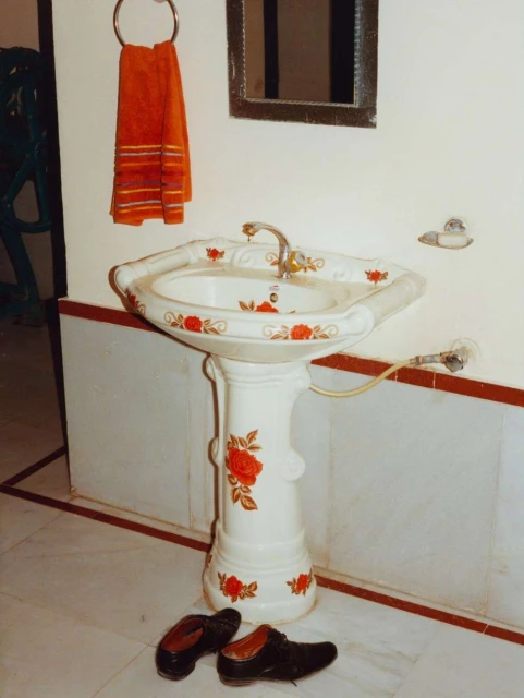 Bathroom interior with a white pedestal sink decorated with red flowers, brown shoes on the floor, orange towel and mirror on the wall.
