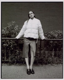 Woman in white puffer jacket leaning against a railing in front of a dark brick wall by photographer Kay Nambiar