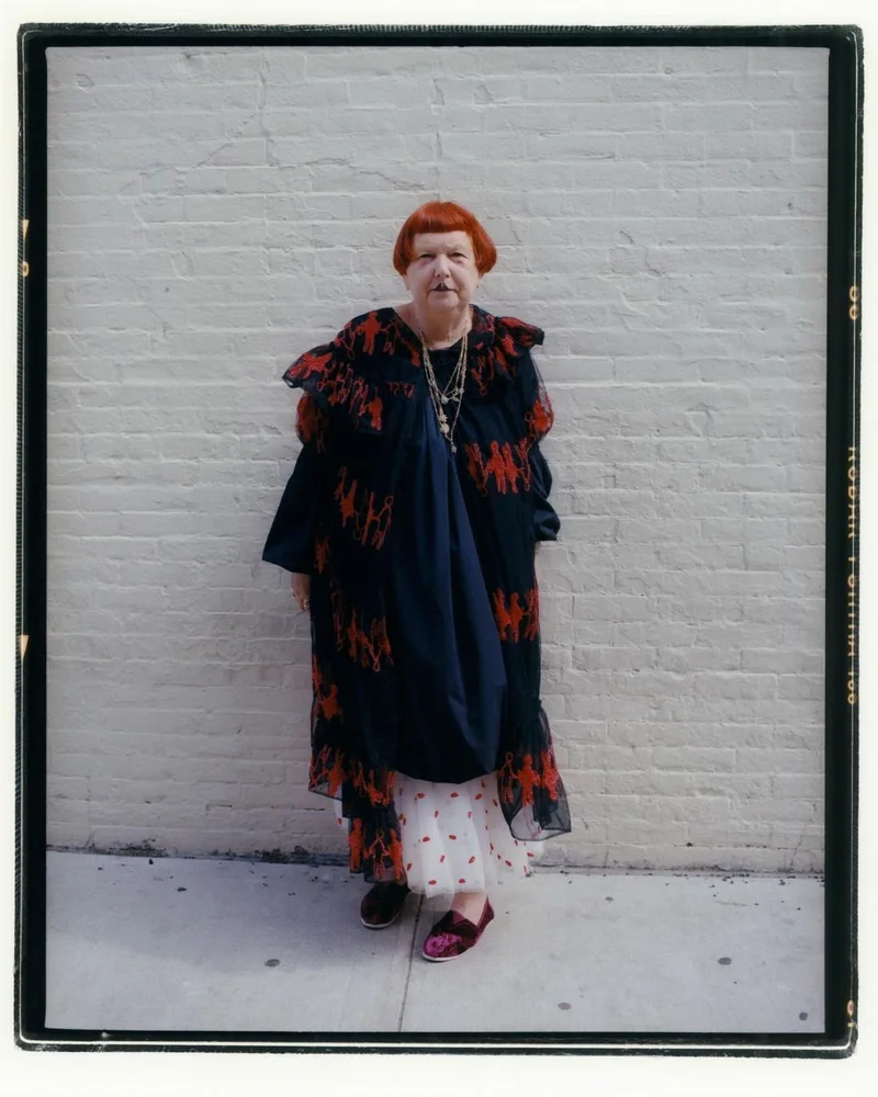 Elderly woman with red hair wearing a dark patterned coat standing against a pale brick wall in Amsterdam