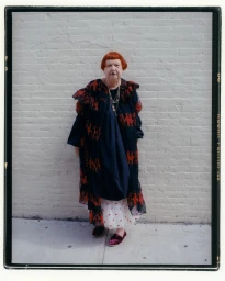 Elderly woman with red hair wearing a dark patterned coat standing against a pale brick wall in Amsterdam