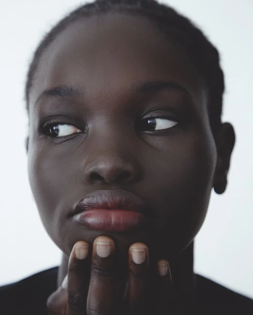 Close-up portrait of a Black woman with dark hair in a black shirt, looking to the left with her hand near her chin in a white setting.