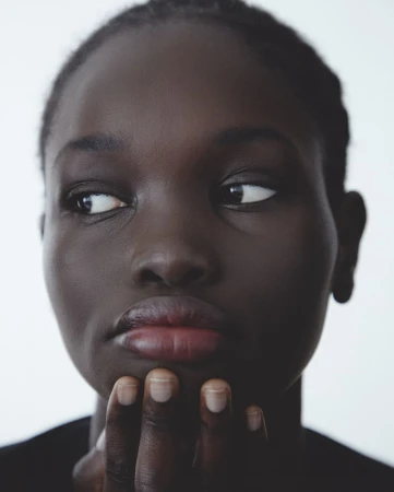 Close-up portrait of a Black woman with dark hair in a black shirt, looking to the left with her hand near her chin in a white setting.