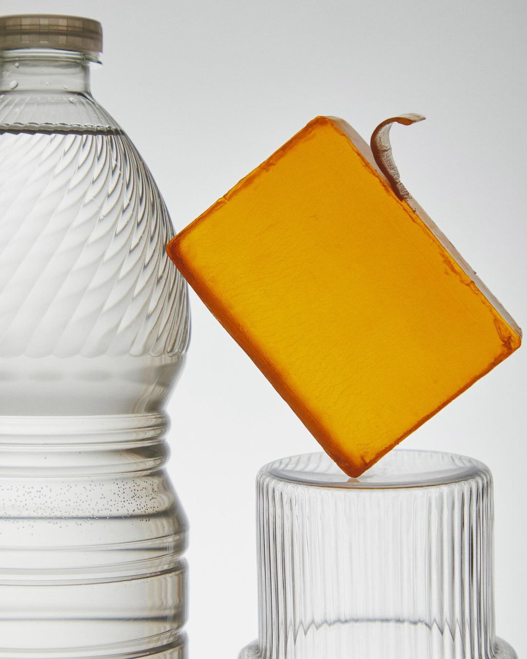 Still life of a block of translucent orange soap balanced on a glass, next to a glass bottle filled with clear liquid on white.
