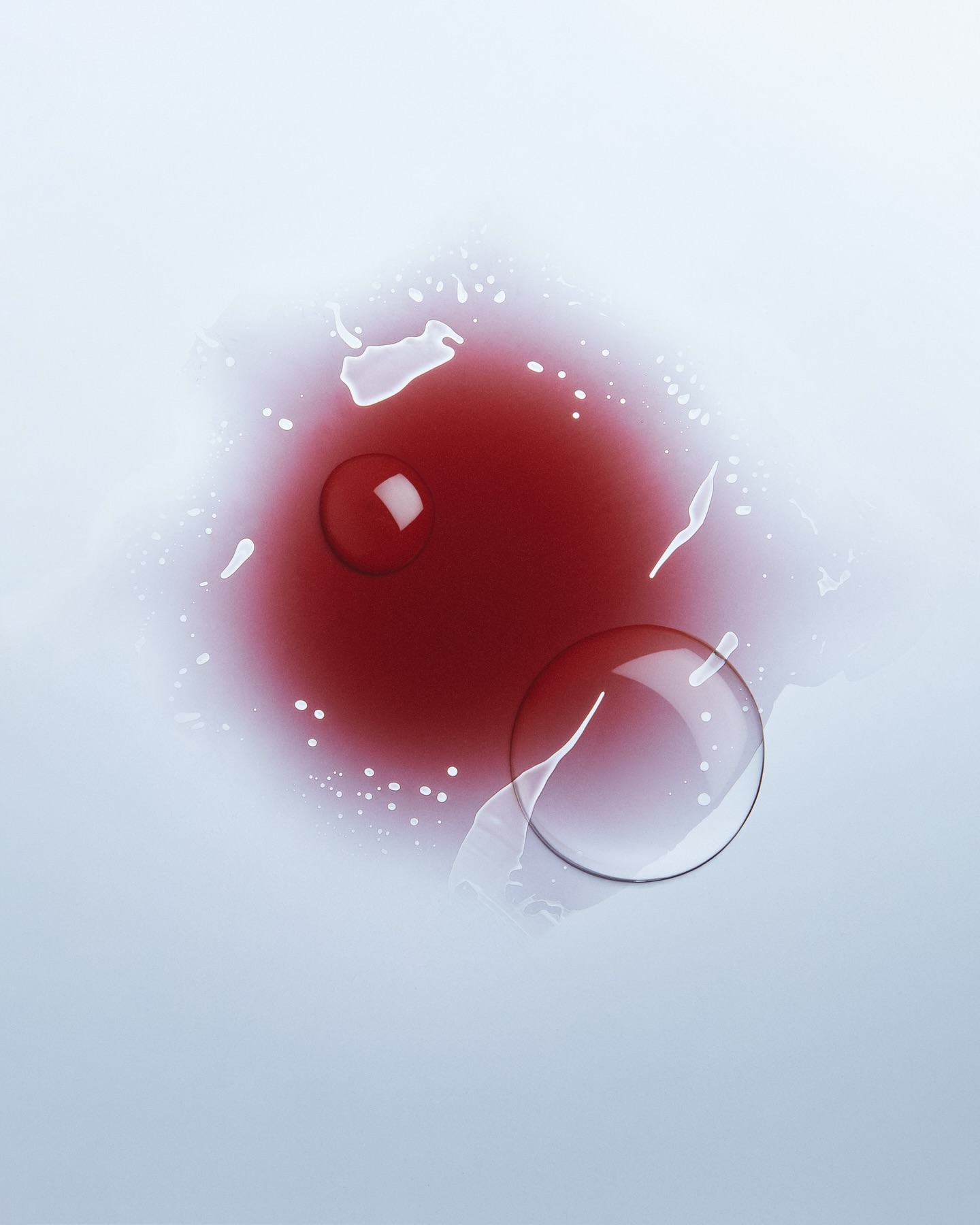 Overhead studio shot of a clear liquid drop and a red liquid pool on a light surface, creating an abstract, minimalist composition.