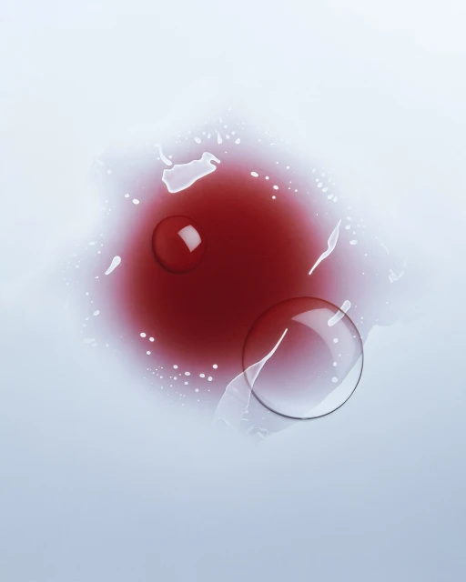 Overhead studio shot of a clear liquid drop and a red liquid pool on a light surface, creating an abstract, minimalist composition.
