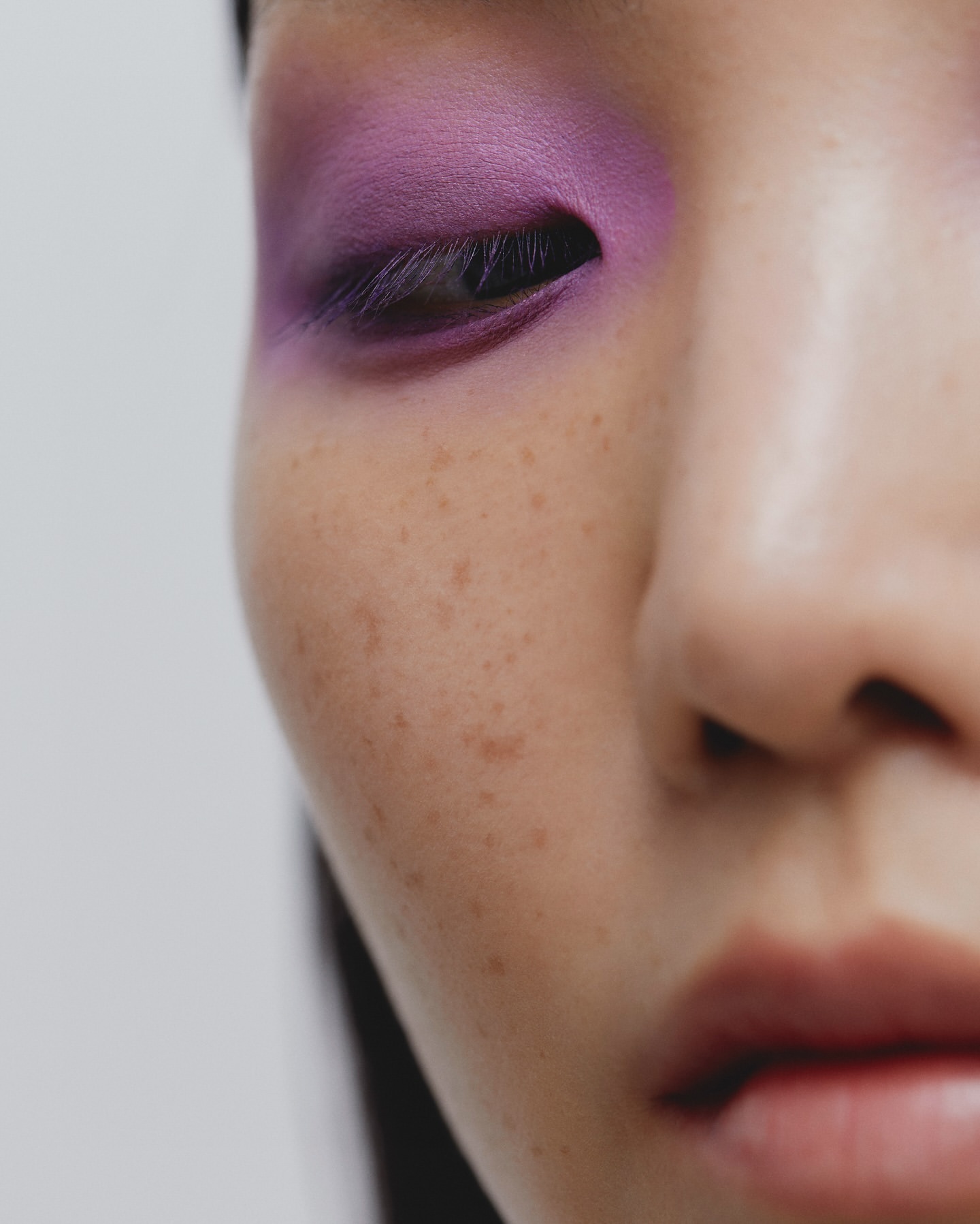 Close-up of a woman's face showcasing vibrant purple eyeshadow, highlighting her eye and skin's texture, against a clean, white backdrop.