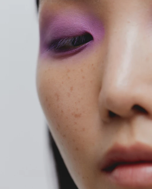 Close-up of a woman's face showcasing vibrant purple eyeshadow, highlighting her eye and skin's texture, against a clean, white backdrop.