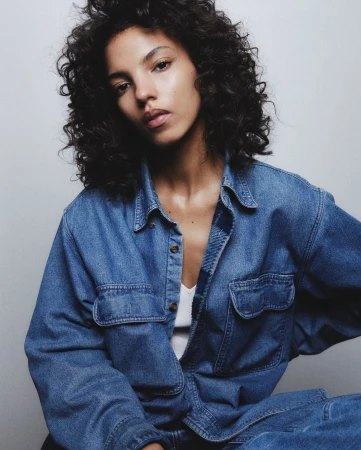 Portrait of a woman with dark curly hair wearing a denim shirt against a white seamless background
