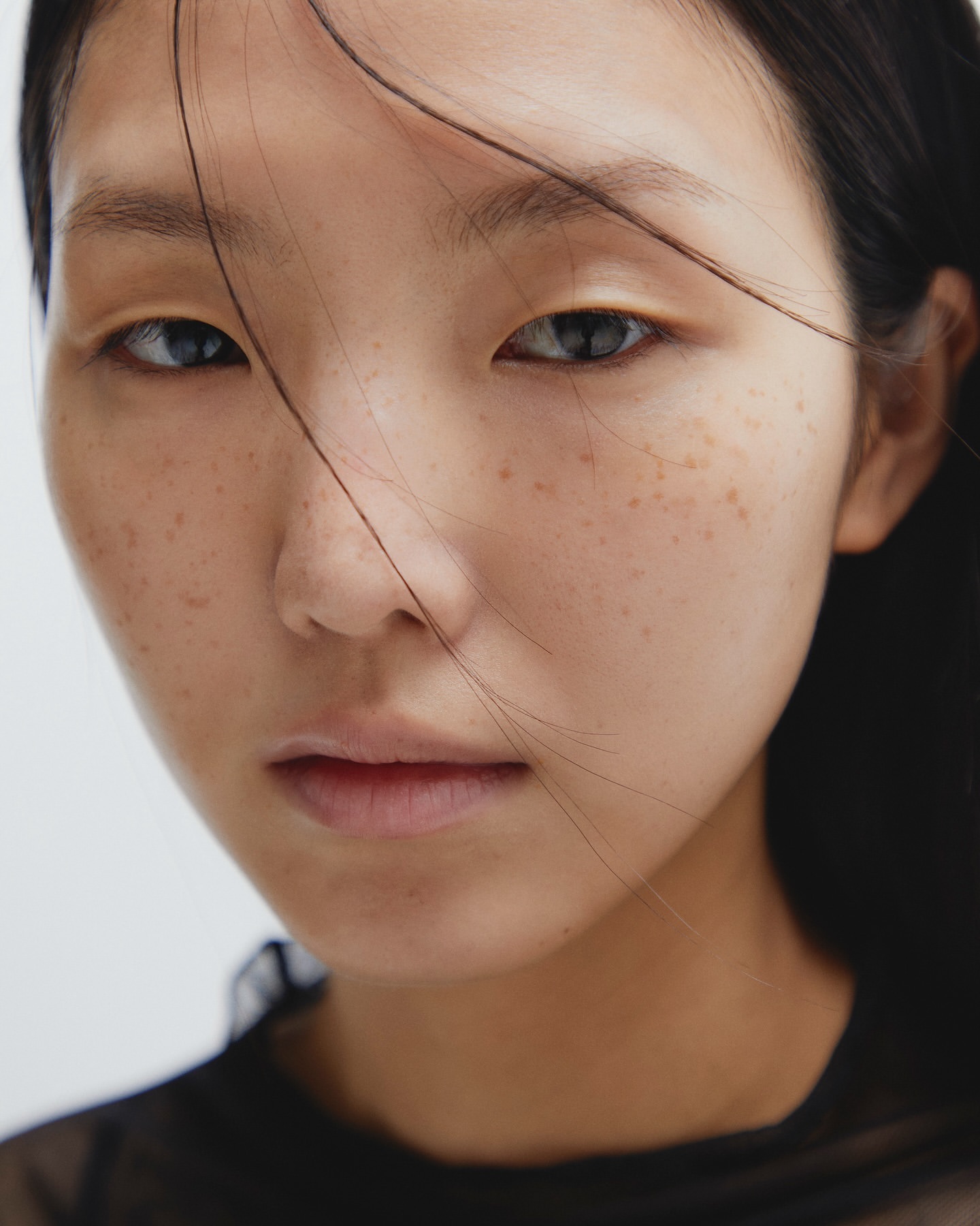 Close-up portrait of a young Asian female model with freckles, light makeup, and loose hair against a neutral background in a beauty context.
