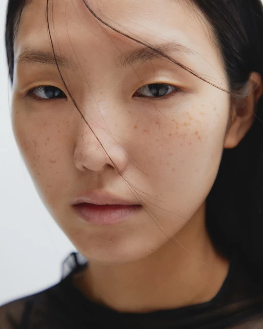 Close-up portrait of a young Asian female model with freckles, light makeup, and loose hair against a neutral background in a beauty context.