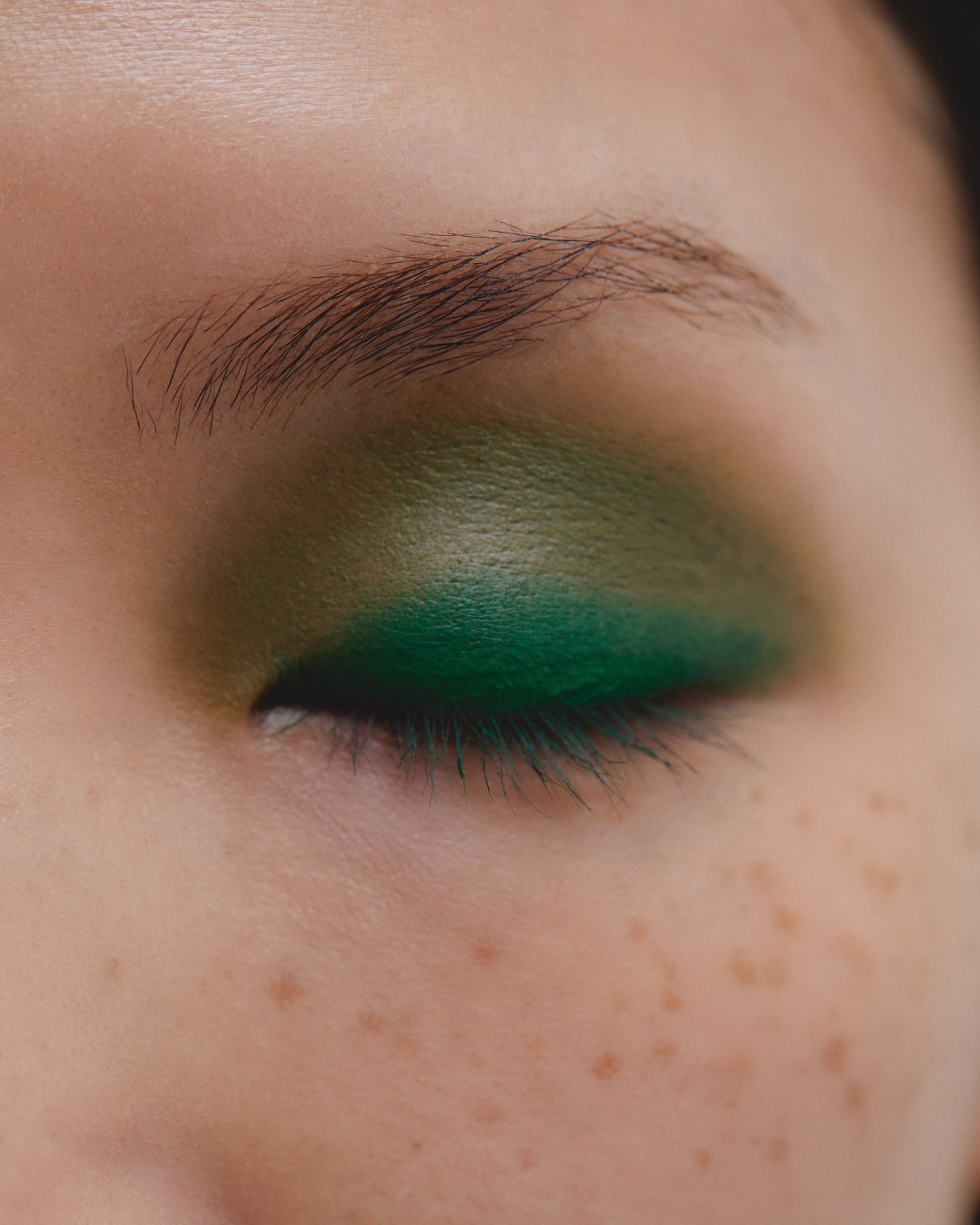 Close-up of a woman's eye with vibrant green eyeshadow. The skin shows visible freckles and natural eyebrow hair.