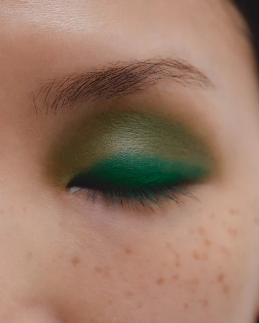 Close-up of a woman's eye with vibrant green eyeshadow. The skin shows visible freckles and natural eyebrow hair.