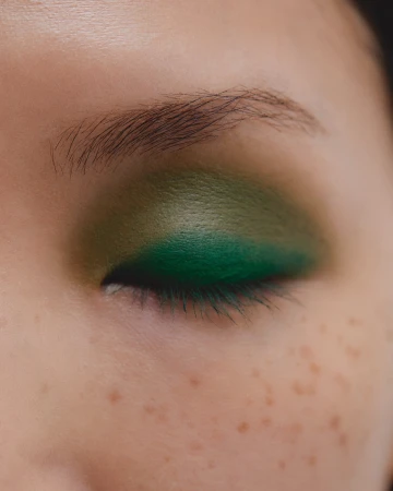 Close-up of a woman's eye with vibrant green eyeshadow. The skin shows visible freckles and natural eyebrow hair.