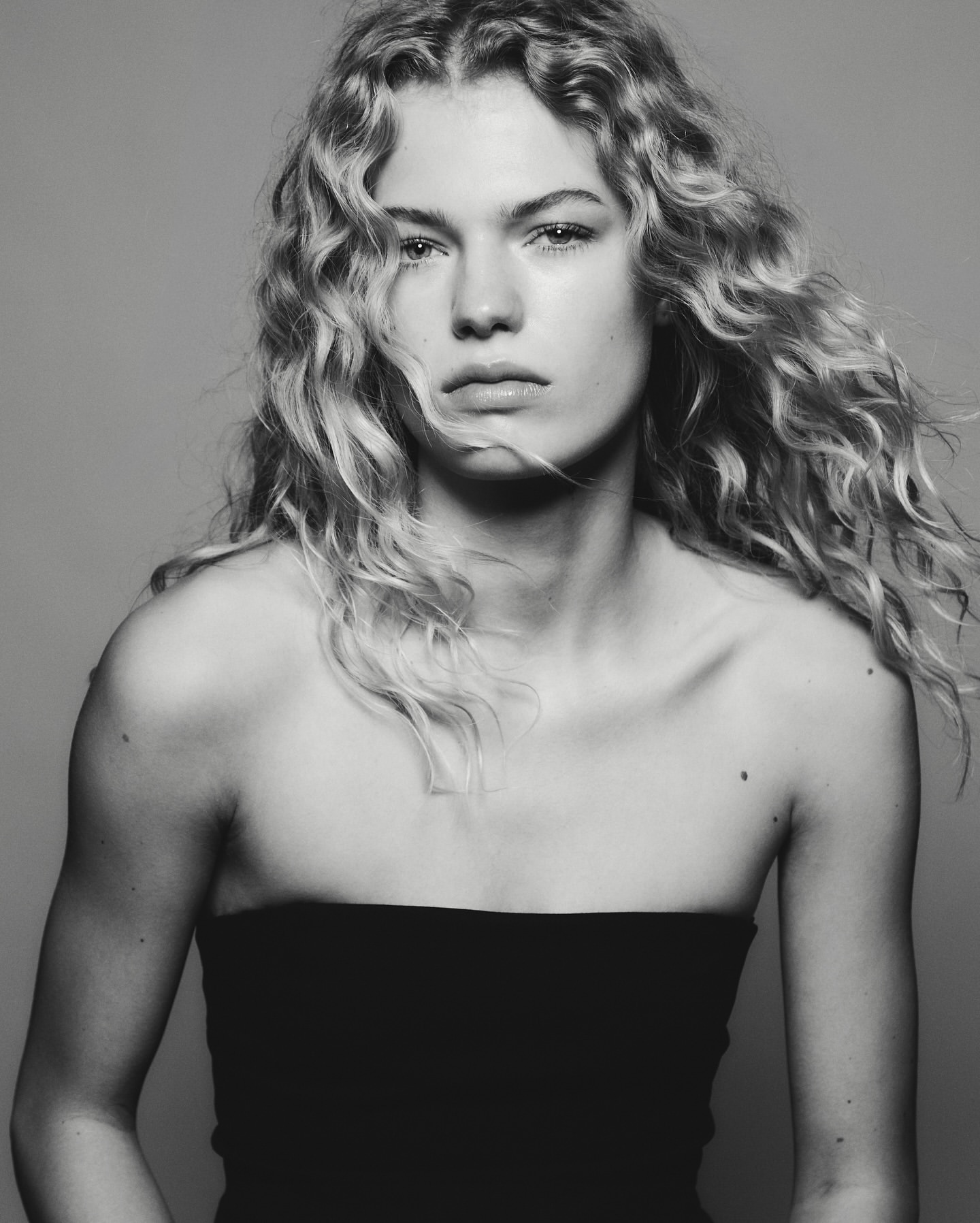 Black and white portrait of a fair-skinned woman with curly blonde hair, wearing a black strapless top against a grey background.