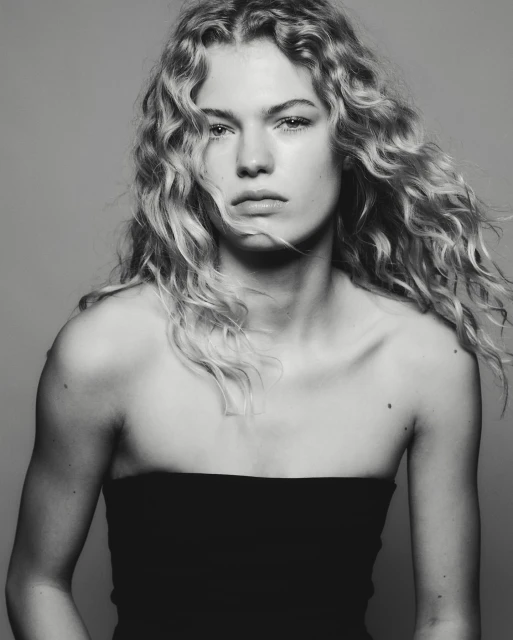 Black and white portrait of a fair-skinned woman with curly blonde hair, wearing a black strapless top against a grey background.
