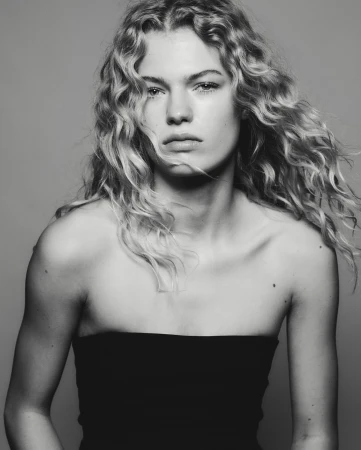 Black and white portrait of a fair-skinned woman with curly blonde hair, wearing a black strapless top against a grey background.