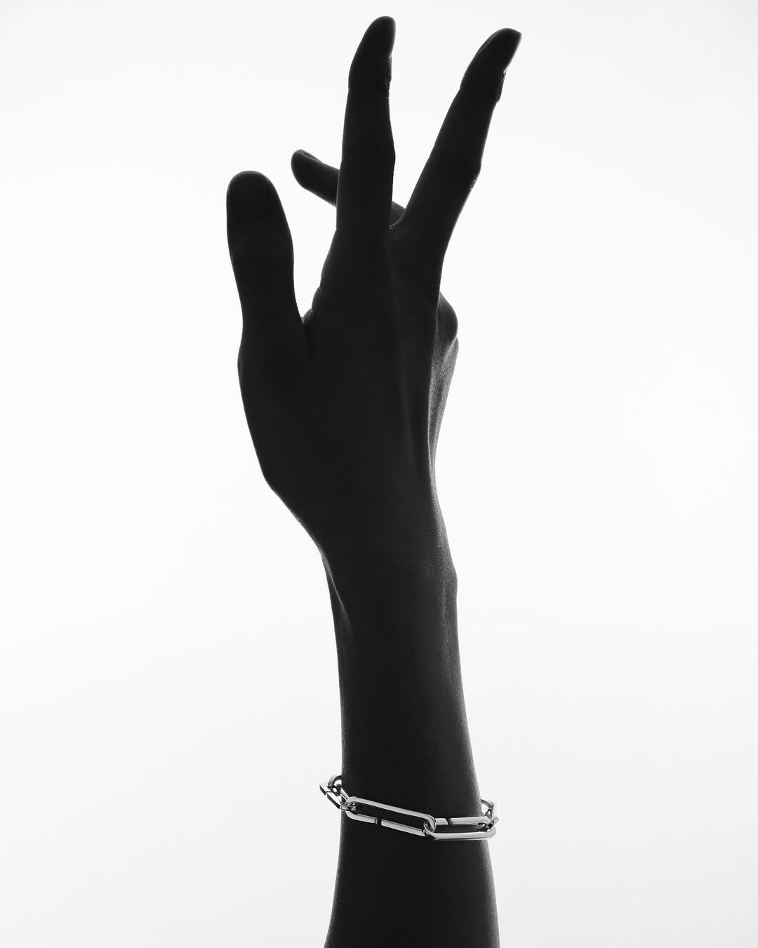 Close-up studio shot of an arm with a silver chain-link bracelet in a high-key, black-and-white image. The hand is raised with fingers extended.