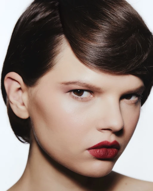 Close-up portrait of an adult female with dark brown, styled hair, wearing red lipstick, and posing in front of a white background.