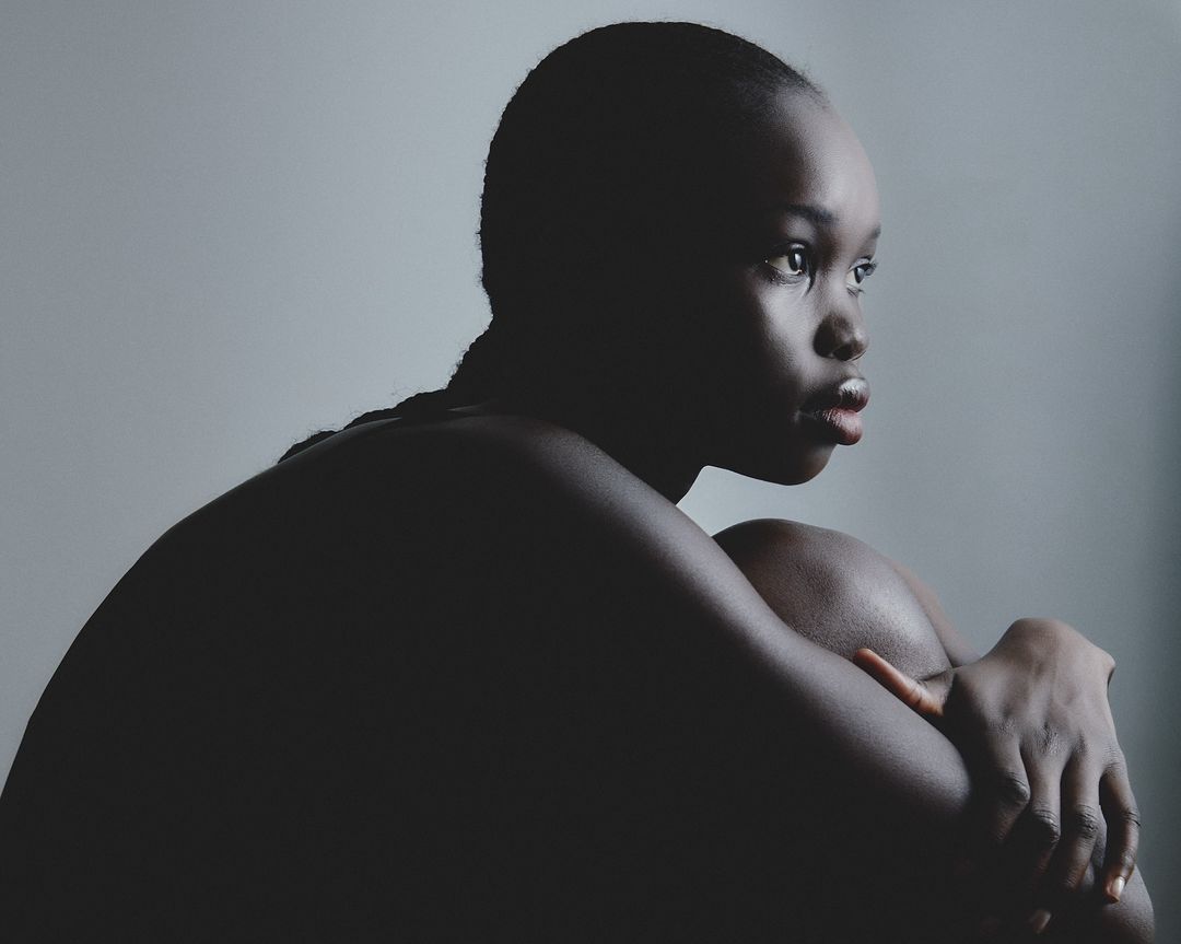 Striking studio portrait of a contemplative Black woman with braided hair, embracing her knees against a gradient grey backdrop.