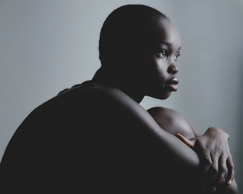 Striking studio portrait of a contemplative Black woman with braided hair, embracing her knees against a gradient grey backdrop.