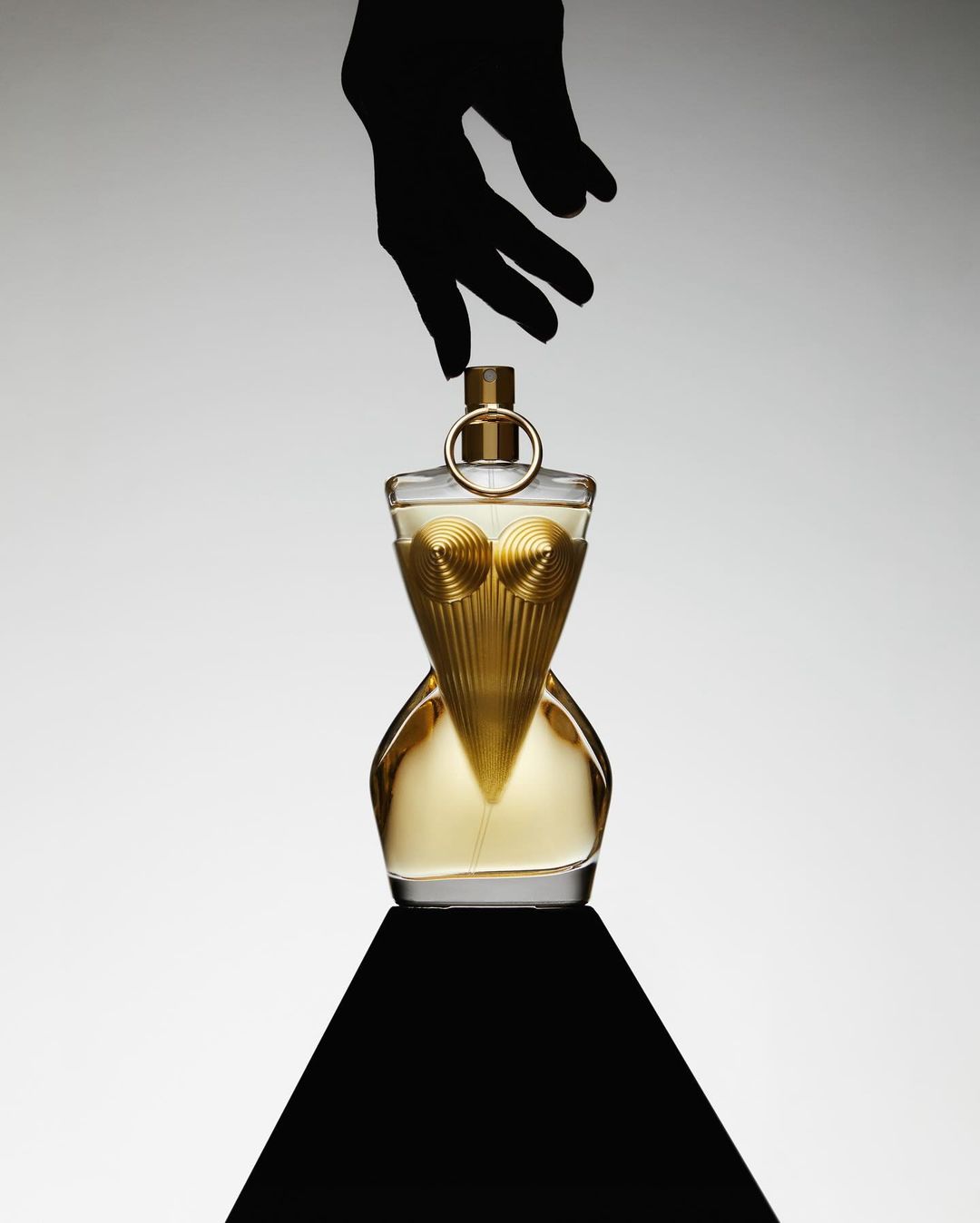 Studio shot of a perfume bottle with gold accents being touched by a hand in a black glove, placed on a black triangular surface.