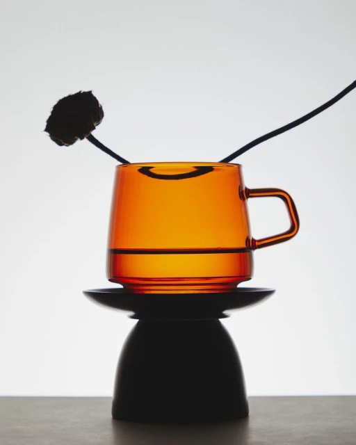 Studio shot of a transparent amber glass mug filled with liquid, resting on a black pedestal with a dark floral stem behind it.