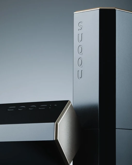 Still life of SUQQU cosmetic containers, showing two dark-colored, geometric packages with gold accents against a gray background.