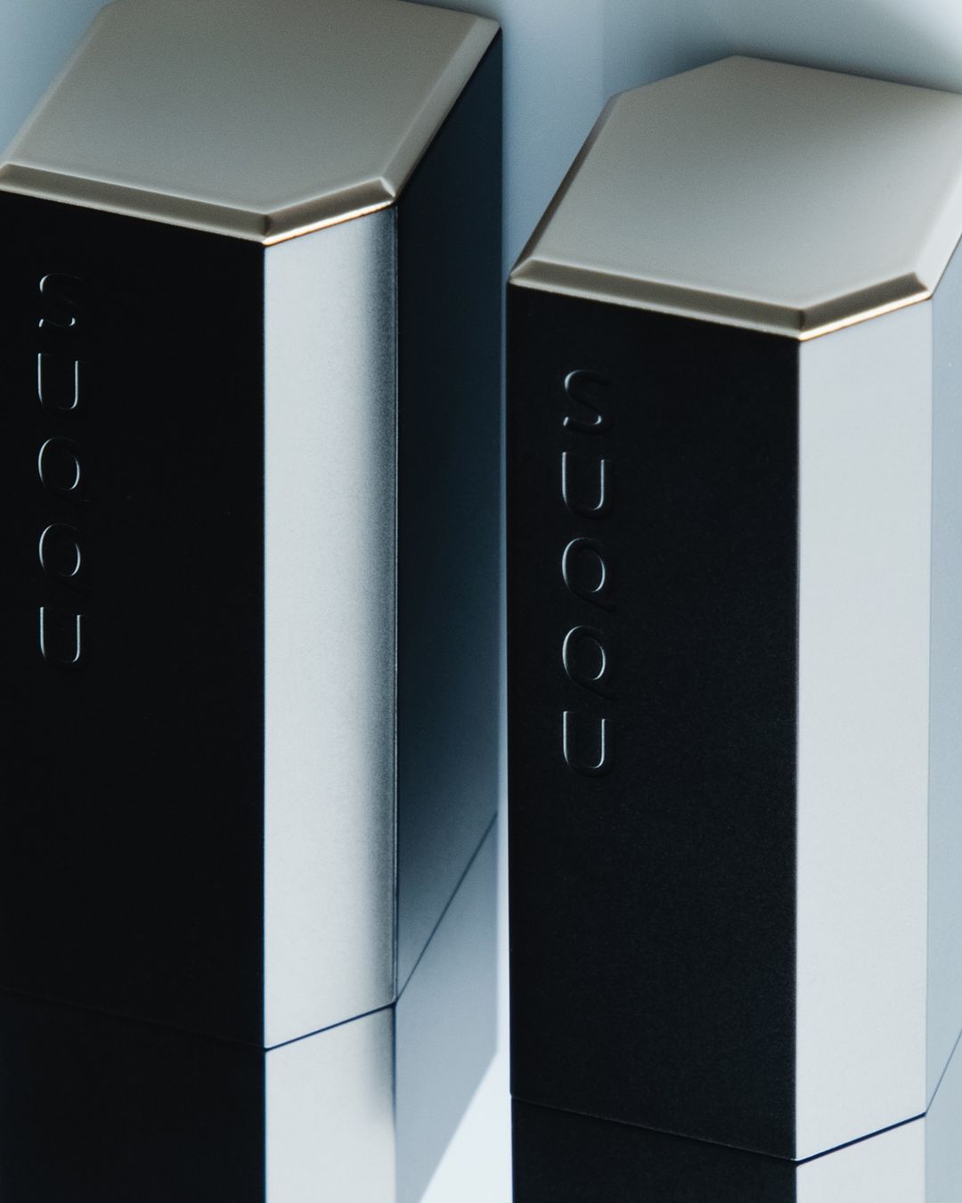 Close-up studio shot of two SUQQU lipstick tubes with black and silver packaging, displaying modern design and brand logo.